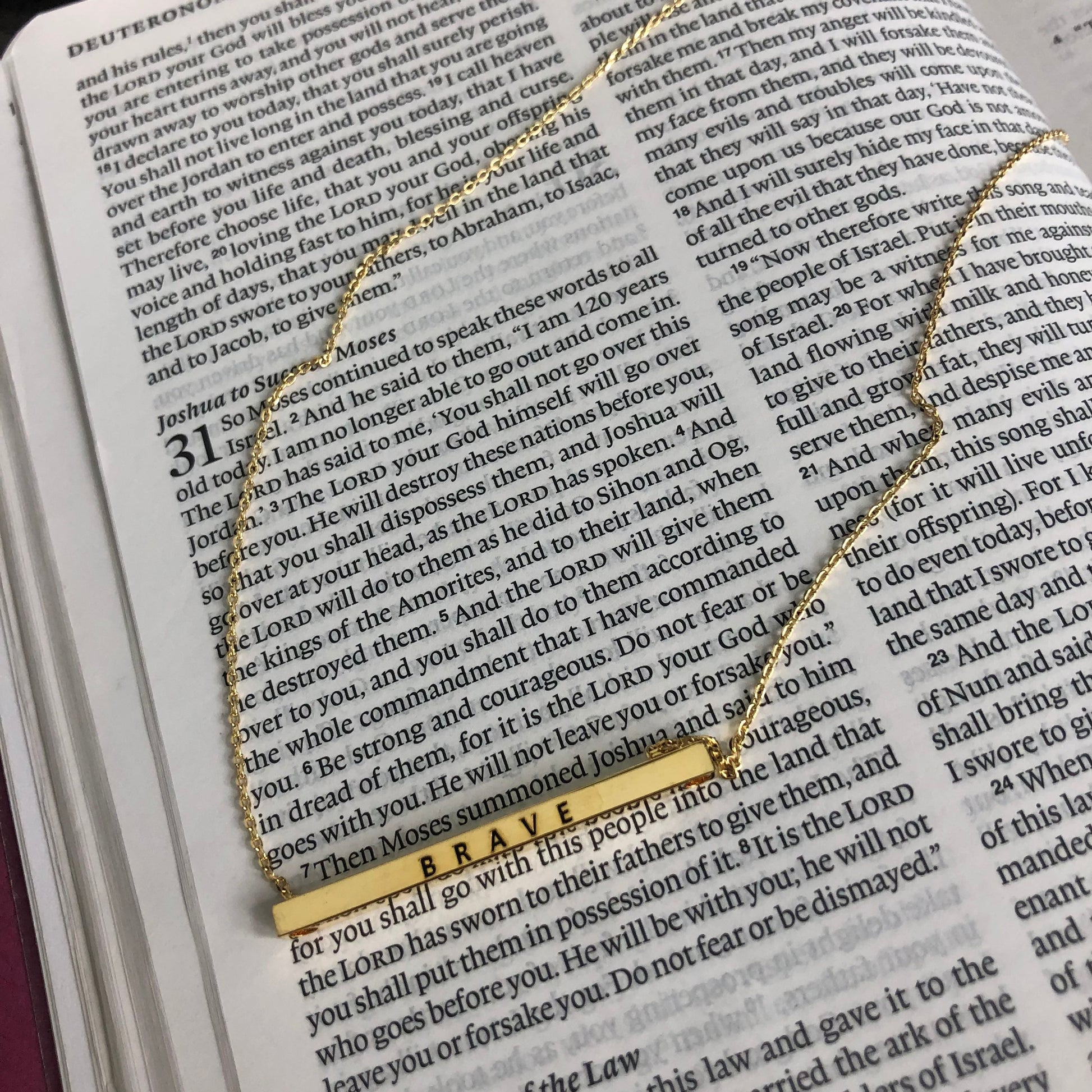 A stylish gold necklace with an engraved rectangular pendant that reads "Brave"